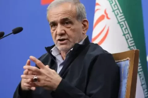 ایرانی صدر مسعود پزشکیان ڈائس پر لگے مائیکروفونز کے سامنے بیٹھے ہوئے گفتگو کر رہے ہیں اور اپنے دونوں ہاتھوں کی انگلیاں آپس میں ملائے اشارہ کر رہے ہیں