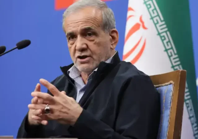ایرانی صدر مسعود پزشکیان ڈائس پر لگے مائیکروفونز کے سامنے بیٹھے ہوئے گفتگو کر رہے ہیں اور اپنے دونوں ہاتھوں کی انگلیاں آپس میں ملائے اشارہ کر رہے ہیں