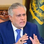 اسحاق ڈار، پاکستانی سیاستدان، ایک لکڑی کی کرسی پر بیٹھے ہوئے بات کر رہے ہیں اور ان کے ہاتھ ہوا میں ہیں۔ انہوں نے نیلے رنگ کا سوٹ اور ٹائی پہنی ہوئی ہے۔ ان کے پیچھے ایک جھنڈا لگا ہوا ہے۔ اسحاق ڈار کے دائیں ہاتھ کی انگلی پر انگوٹھی ہے۔