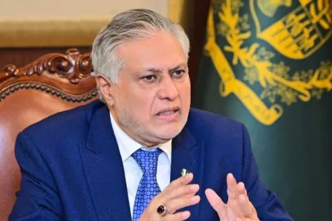 اسحاق ڈار، پاکستانی سیاستدان، ایک لکڑی کی کرسی پر بیٹھے ہوئے بات کر رہے ہیں اور ان کے ہاتھ ہوا میں ہیں۔ انہوں نے نیلے رنگ کا سوٹ اور ٹائی پہنی ہوئی ہے۔ ان کے پیچھے ایک جھنڈا لگا ہوا ہے۔ اسحاق ڈار کے دائیں ہاتھ کی انگلی پر انگوٹھی ہے۔